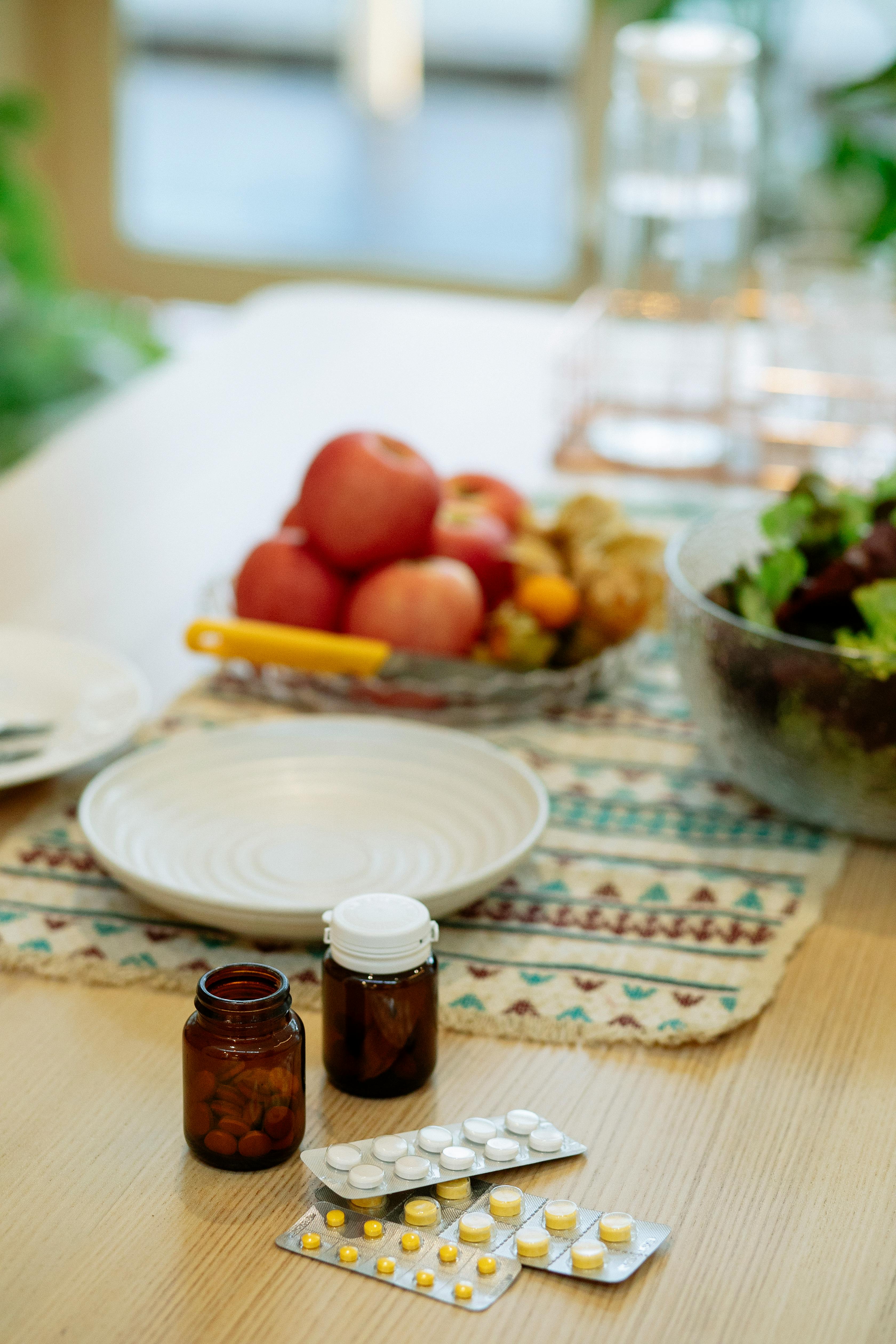 bottle and stips of medicine on a table with an empty plate and another plate filled with red apples in the background